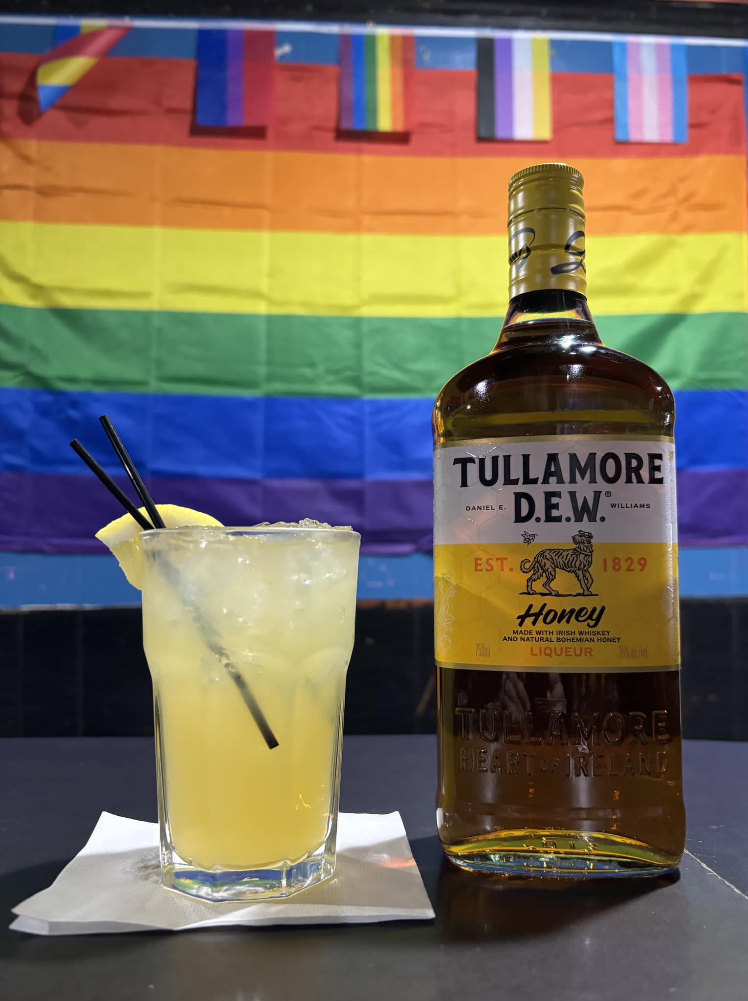 a Tully Honey Sour with a lemon wedge and a bottle of Tullamore Dew honey with pride flags in the background