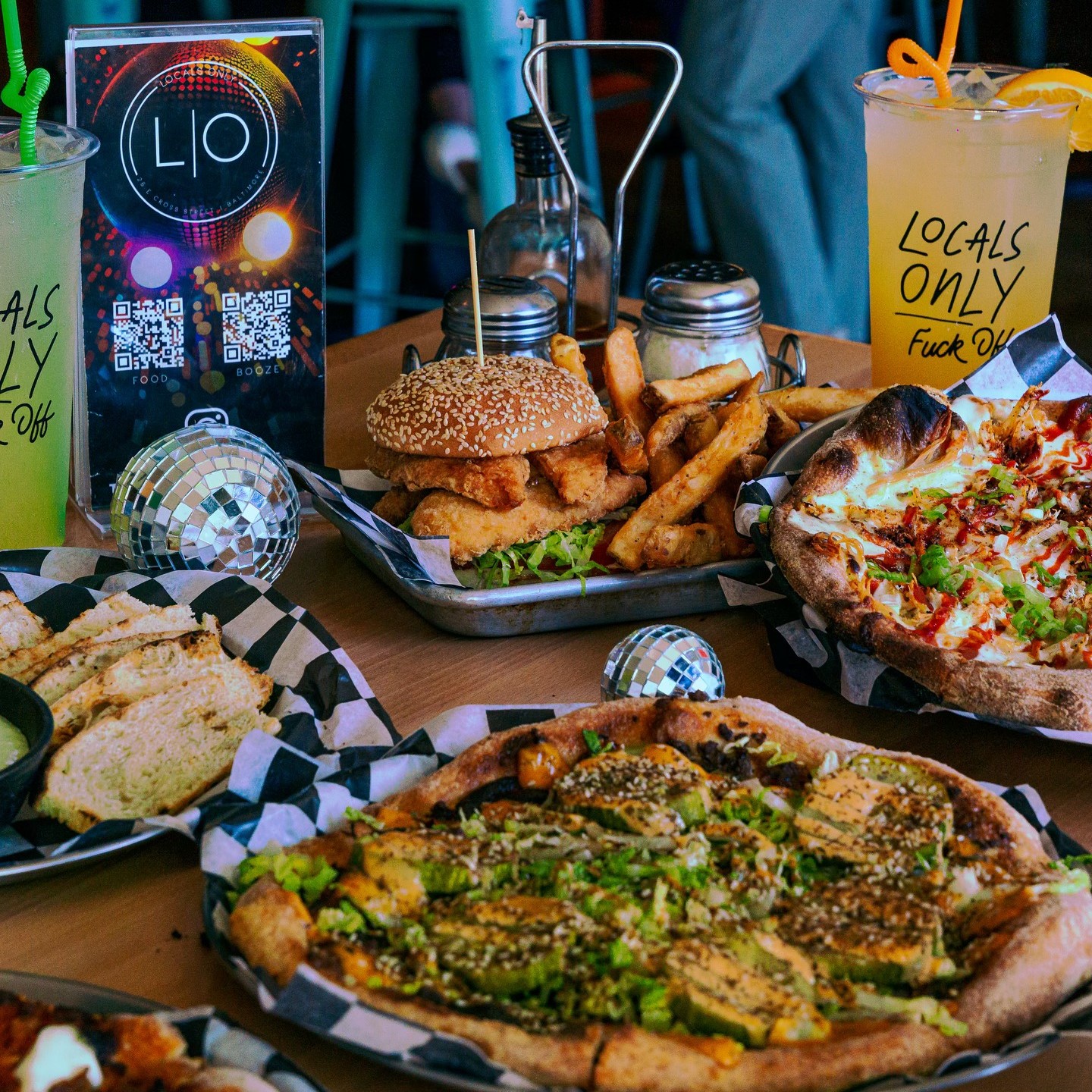 a table with two drinks and several platters of food, including pizzas and a nugget sandwich with fries