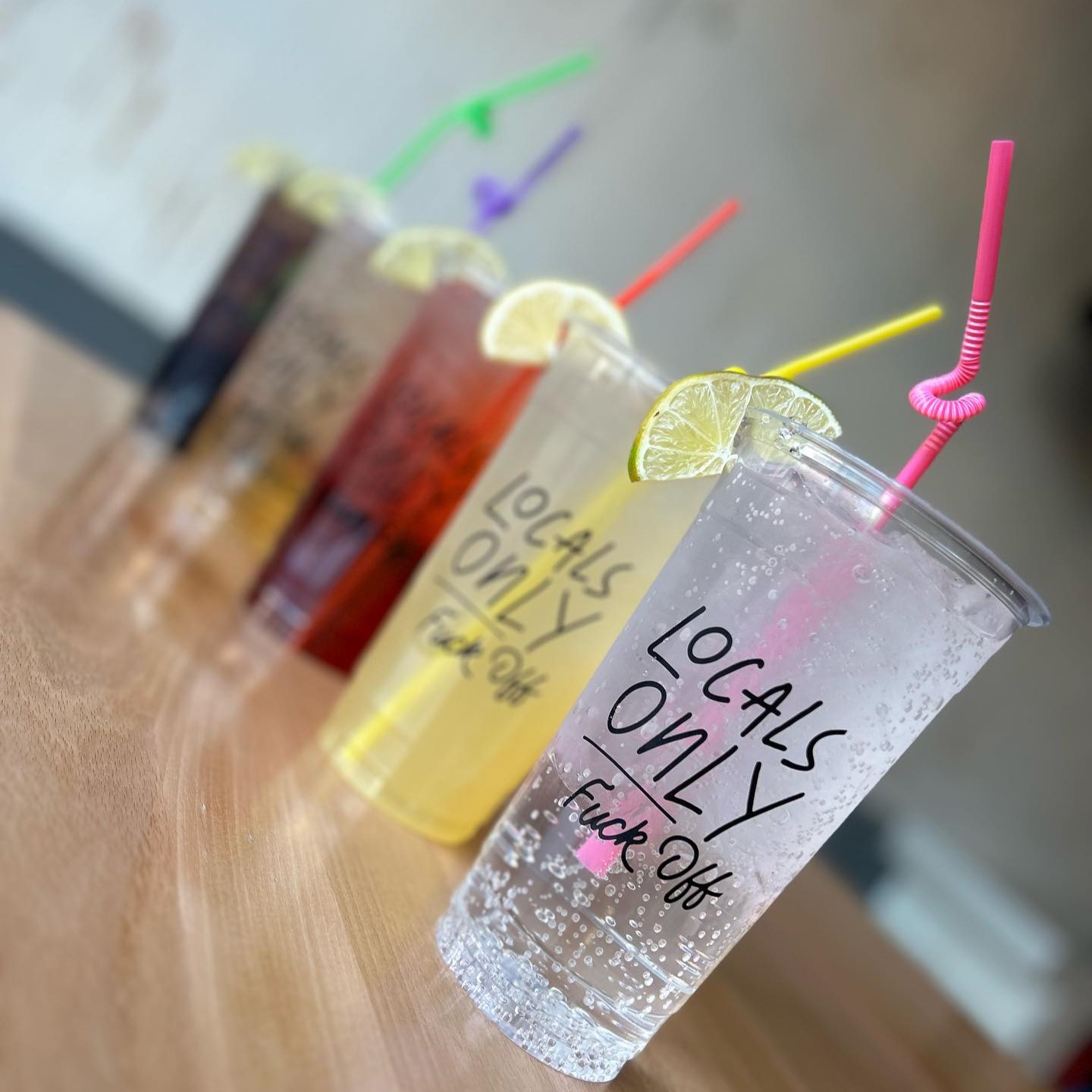 a row of mixed drinks of various colors in locals only glasses with colorful bendy straws and citrus wedges