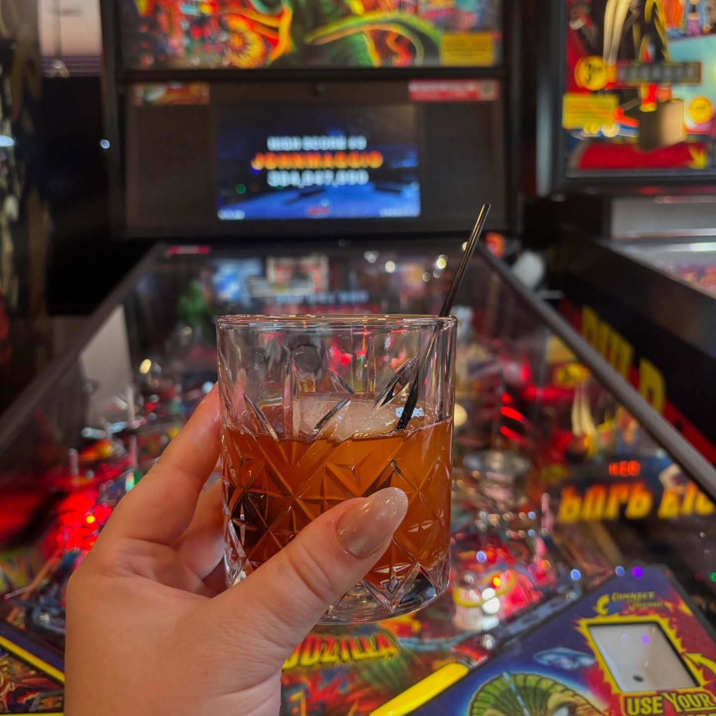 a hand holding a s'mores old fashioned in front of a pin ball machine